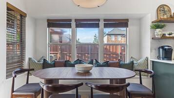 Un coin repas avec une table ronde et six chaises, de grandes fenêtres équipées de stores et une vue sur une terrasse en bois et la maison.