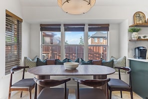 A dining area with a round table and six chairs, large windows with blinds, and a view of a wooden deck and house.