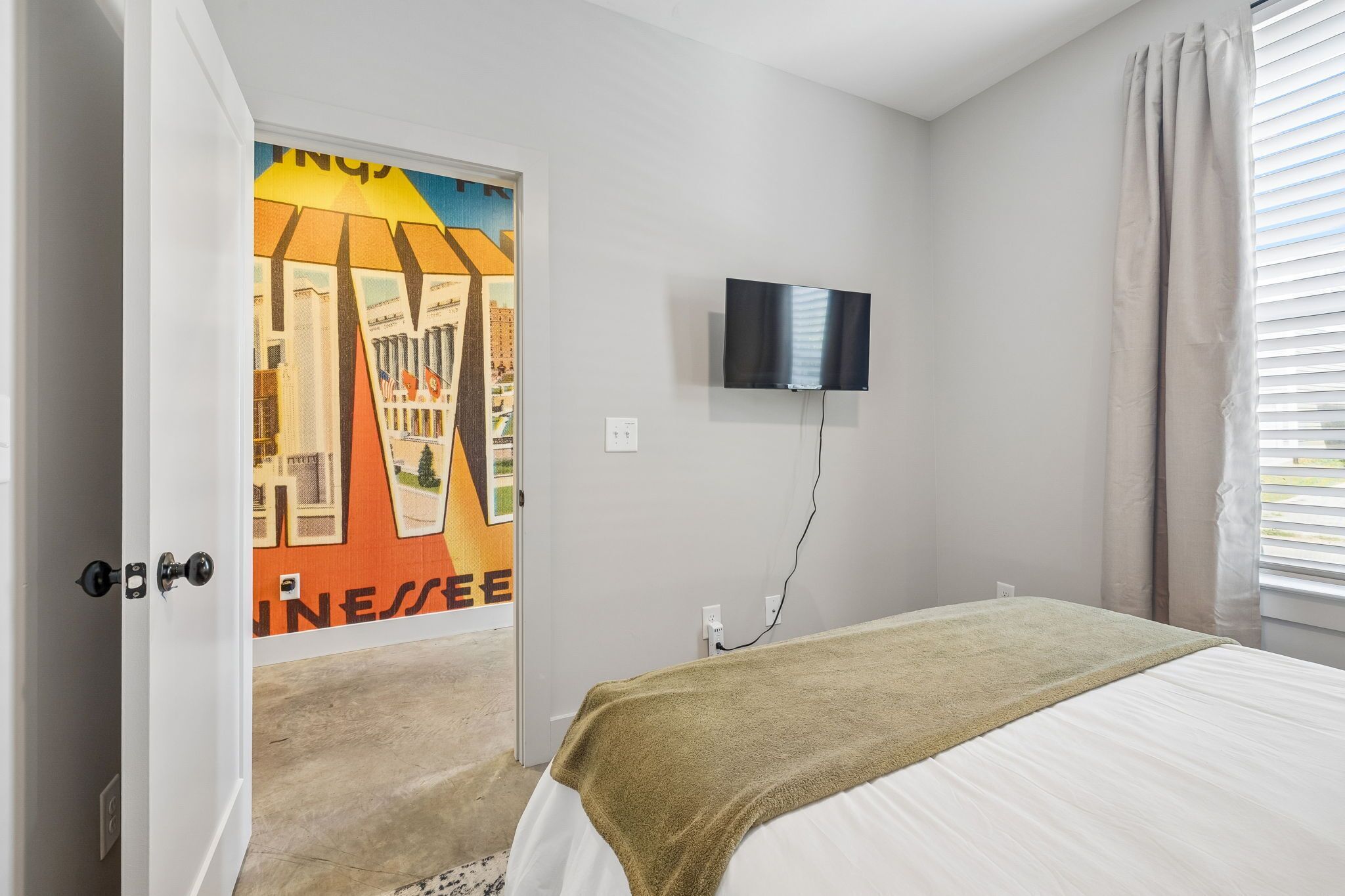 Un dormitorio con una cama, un televisor montado en la pared y una puerta con un mural colorido.