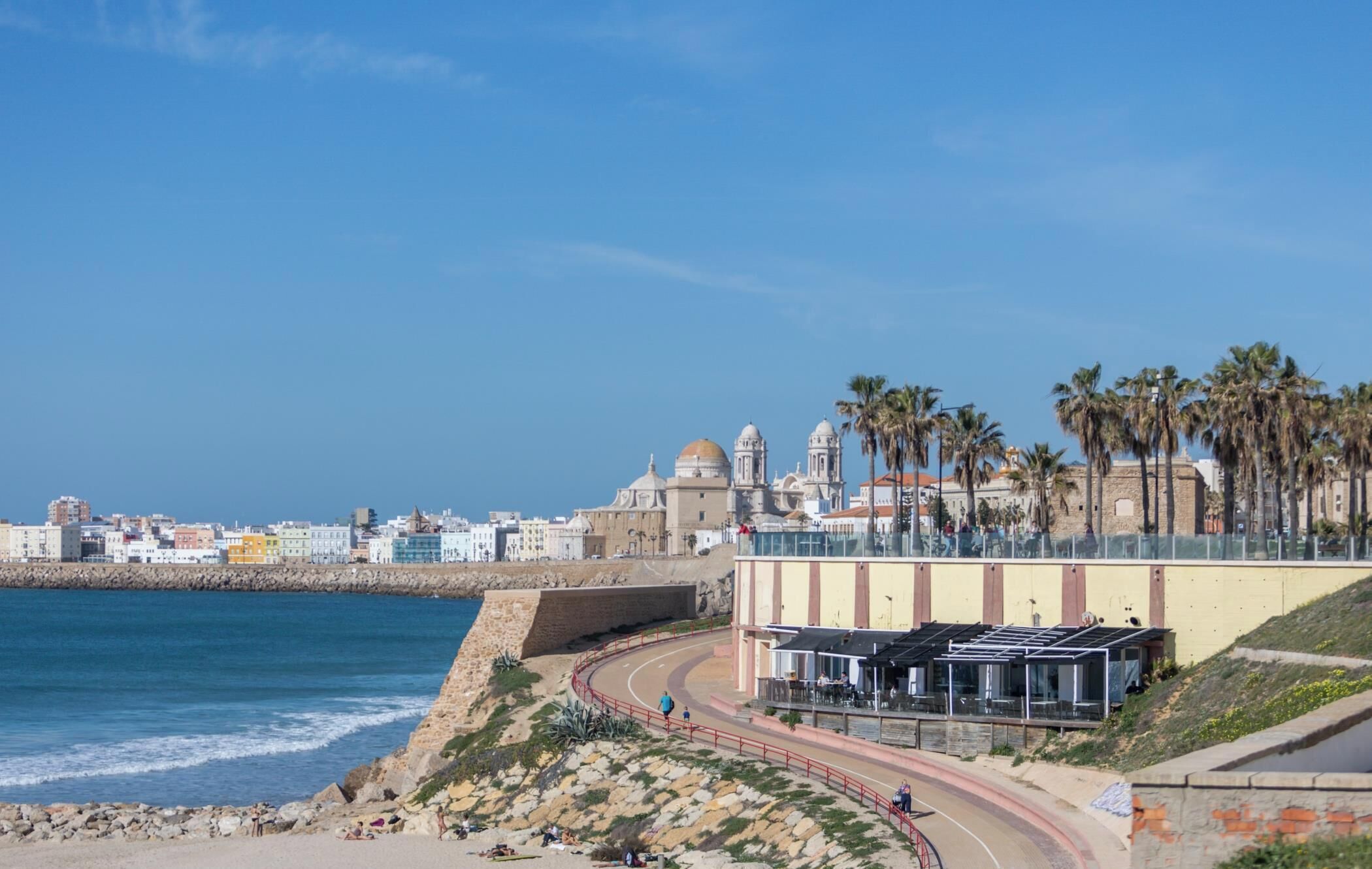 A coastal cityscape with a prominent historical building, a curved road, and a beach area.