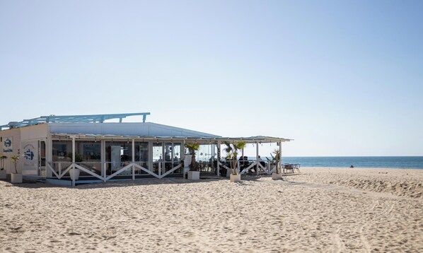 Ein Gebäude am Strand mit überdachter Terrasse, freiem Blick auf den Ozean und einem sandigen Strand im Vordergrund.