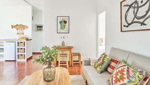 A modern living room with a sofa, colorful cushions, a wooden coffee table, a vase with greenery, a hanging light fixture, a kitchen area with a fish-shaped lamp, and a framed abstract painting on the wall. - New penthouse with a private terrace in the heart of Cádiz. (Ciudad de Cadiz)