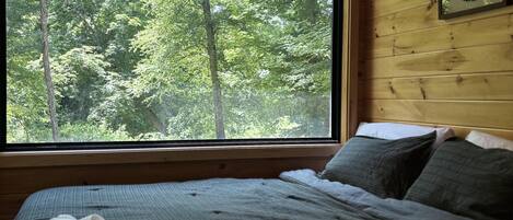 A wooden cabin bedroom with a large window, a bed with pillows, and a framed picture on the wall.