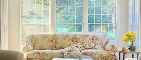 A room with a wooden ceiling, a floral sofa, a matching ottoman, and a view of greenery through large windows.