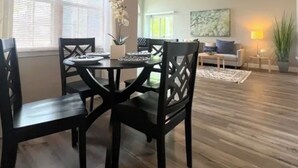 Ein Essbereich mit Tisch und Stühlen, ein Wohnzimmer mit Sofa und Lampe sowie ein großes Fenster mit Jalousien.