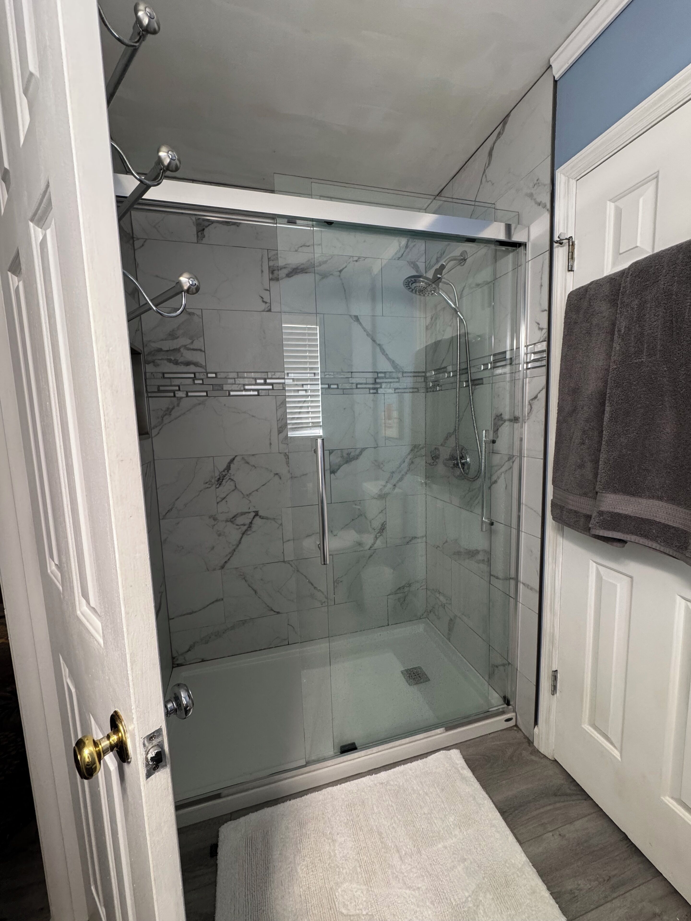 A glass-enclosed shower with a marble wall, a towel rack, and a white door.