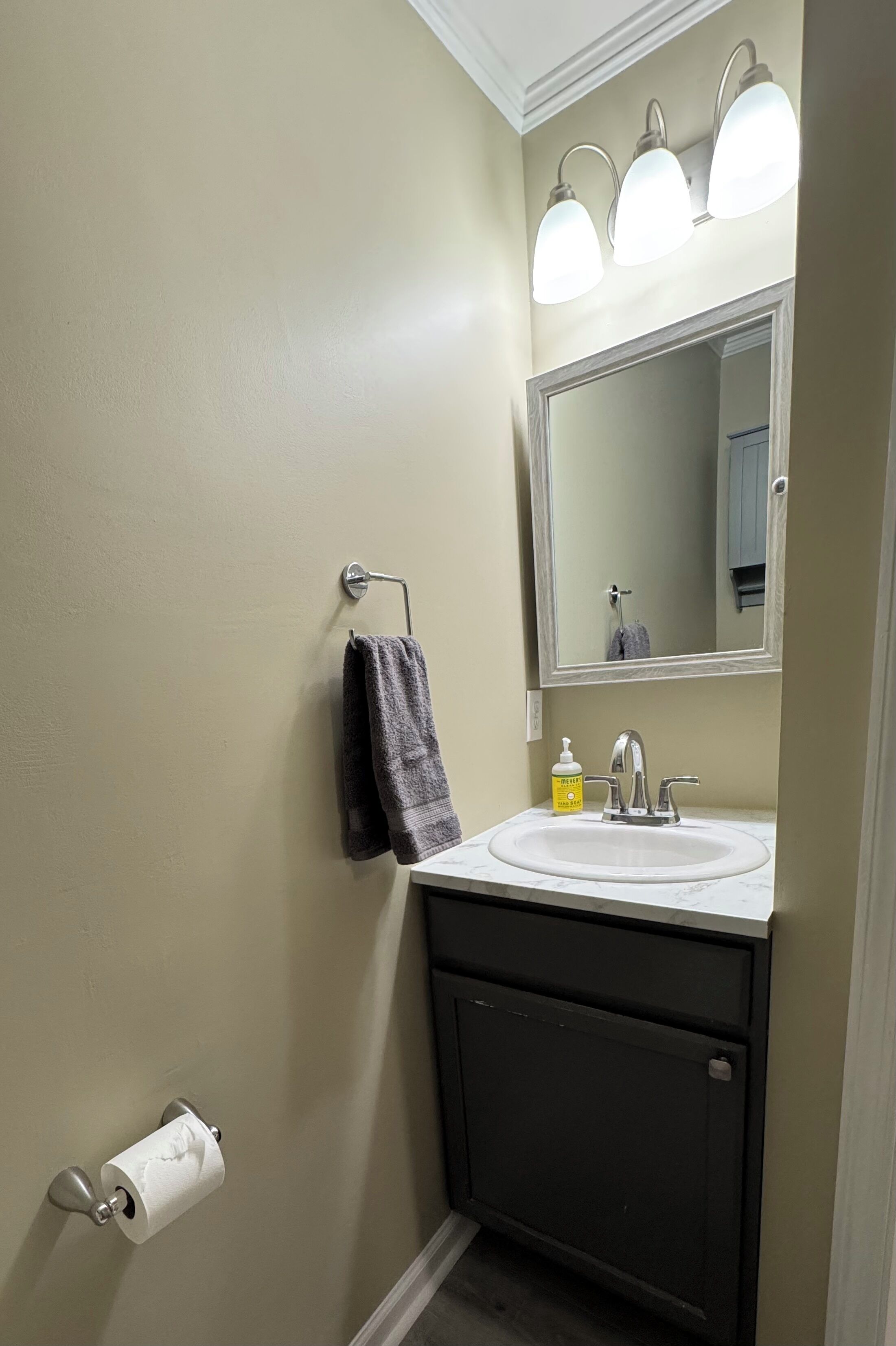 A bathroom with a sink, mirror, towel rack, toilet paper, and a soap dispenser.