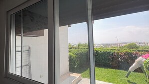Een kamer met een groot raam, een witte muur en uitzicht op een groen gazon en een gebouw in de verte.