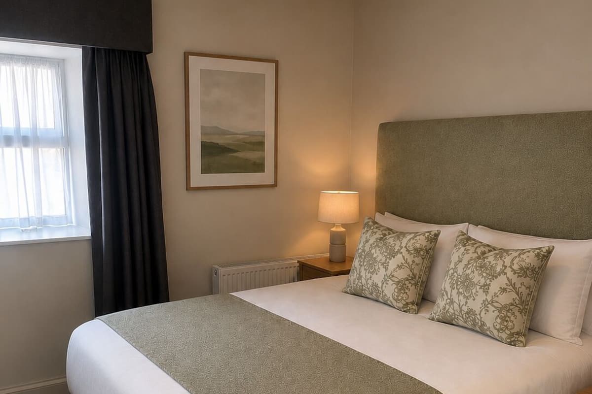 Habitación de hotel con una cama grande, una mesita de noche con lámpara, un cuadro enmarcado en la pared y una ventana con cortinas.