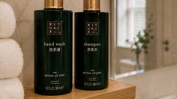 Two bottles of Ritual of Jing hand wash and shampoo on a wooden shelf in a bathroom with a marble wall and a large mirror.