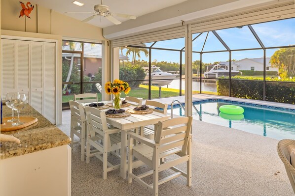 A dining area with a table set for four, a pool, and a view of boats and greenery. - Sunsets & Serenity: Waterfront Canal Home w/ Saltwater Pool in Punta Gorda Isles (Punta Gorda)