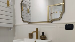 A bathroom with a white sink, gold fixtures, a mirror with a gold frame, a wooden shelf, and a black trash bin. - CIATTONI GUEST HOUSE STUDIO SUITE (Pescara)