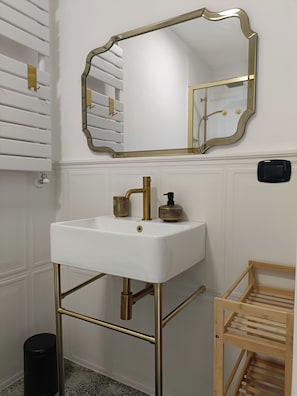 A bathroom with a white sink, gold fixtures, a mirror with a gold frame, a wooden shelf, and a black trash bin. - CIATTONI GUEST HOUSE STUDIO SUITE (Pescara)