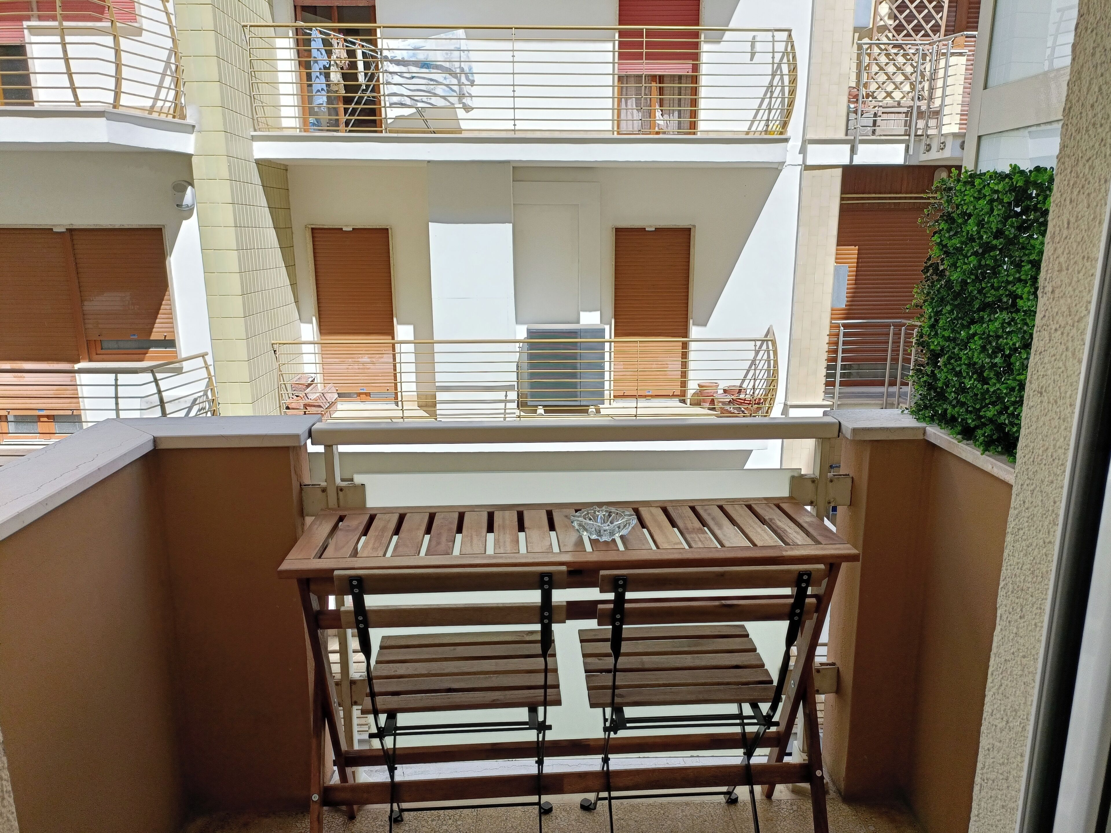 A balcony with a wooden table and bench, a view of the building's interior, and a green hedge.