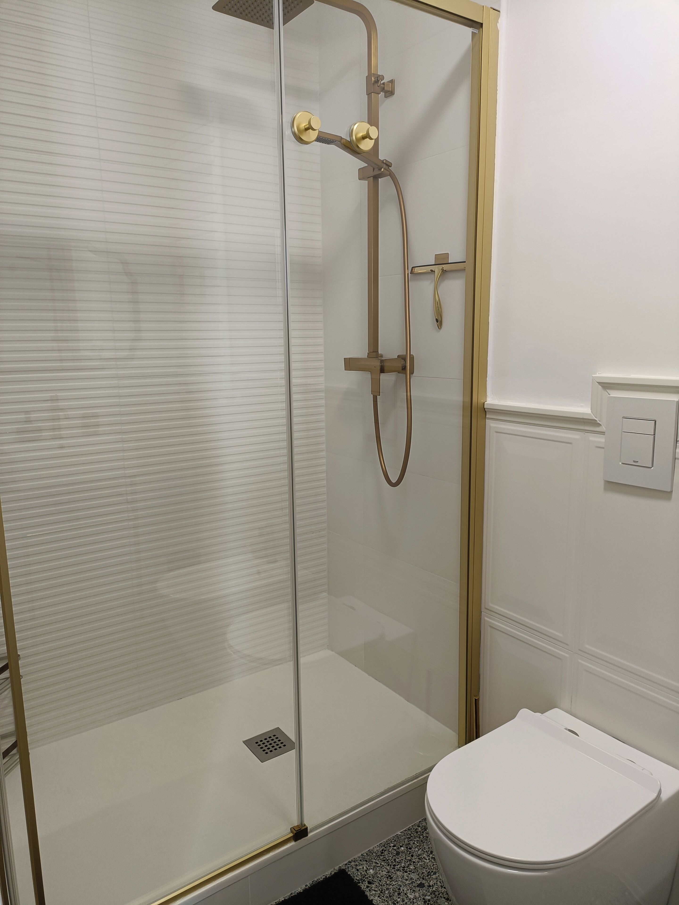 Eine gläserne Dusche mit goldenem Wasserhahn und einer weißen Toilette in einem Badezimmer mit weißen Wänden und Fliesenboden.