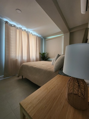 Ein Schlafzimmer mit einem Bett, einem Nachttisch mit Lampe, einer Topfpflanze und einem Fenster mit Vorhängen.
