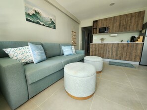 Ein Wohnzimmer mit einer Couch, Poufs, einer Küchenzeile mit Holzschränken, einer Mikrowelle und einem Wellenbild an der Wand.