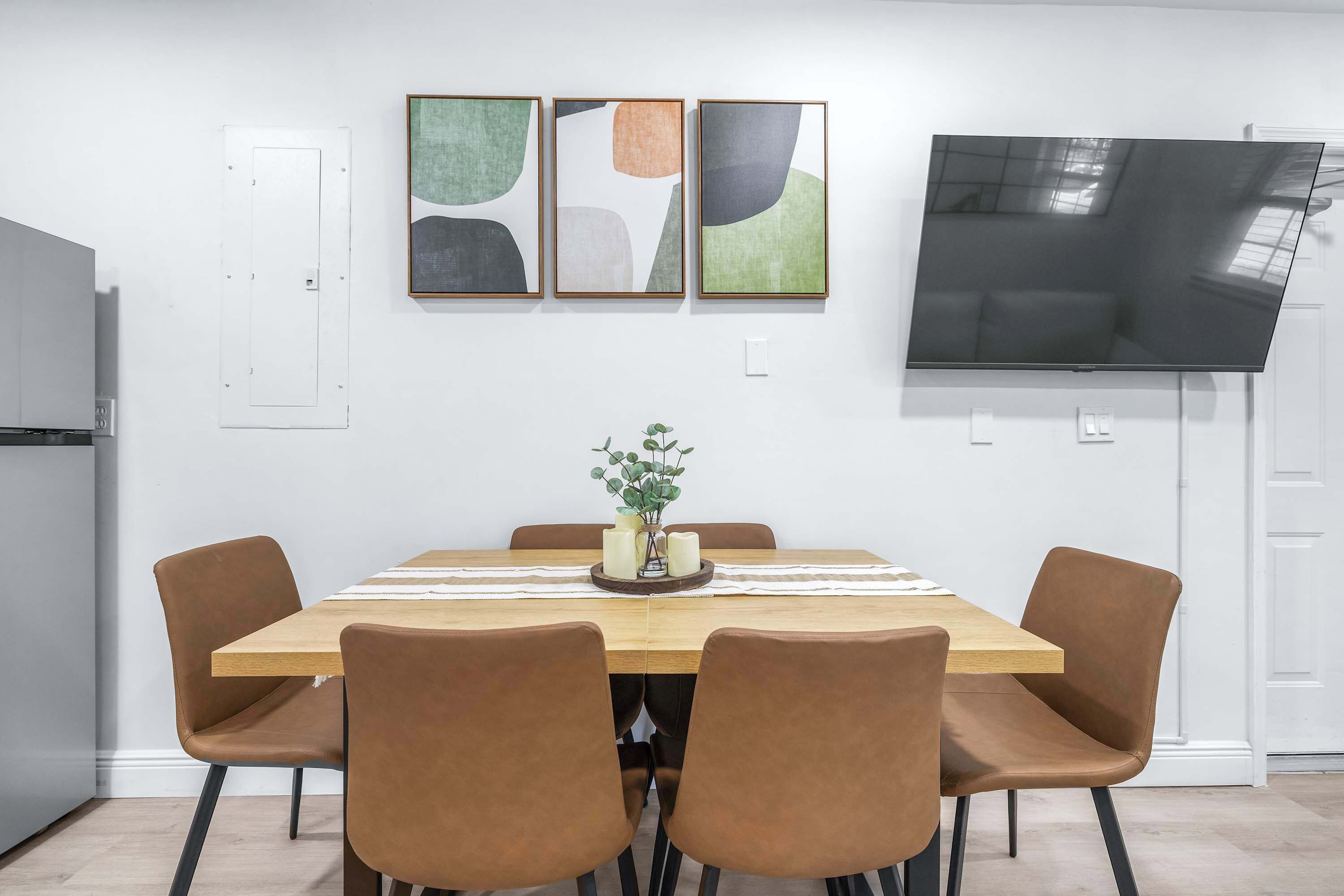 Un coin repas avec une table en bois, six chaises marron, un vase avec des bougies et une œuvre d’art abstraite accrochée au mur.