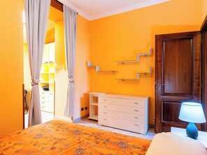 A bedroom with an orange wall, a wooden door, a bedside table with a lamp, a dresser, and a patterned bedspread. - Apartment Daisy by Interhome (Imperia)