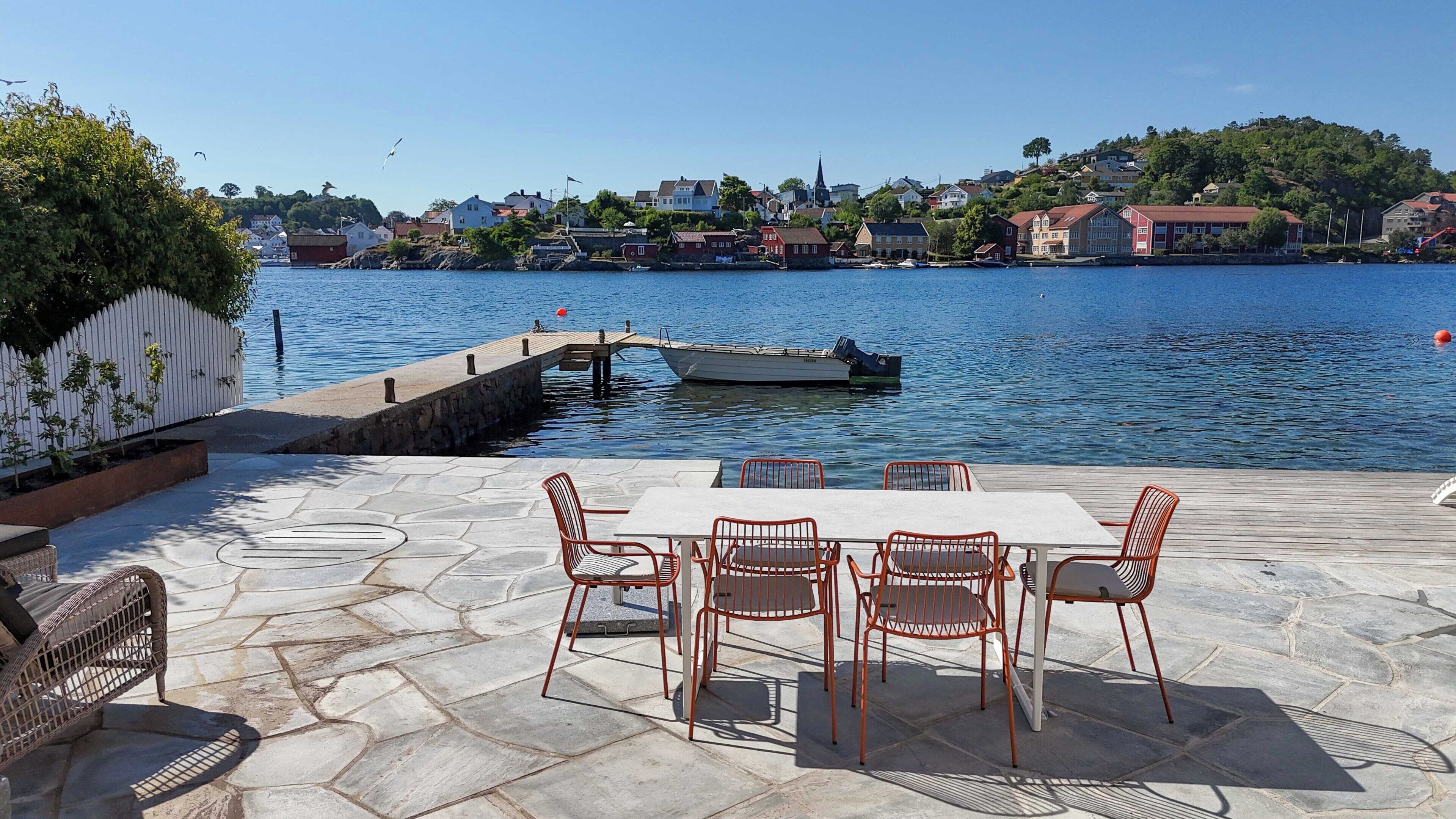A waterfront patio with stone flooring, a table with chairs, and a view of the water with boats and buildings in the background.