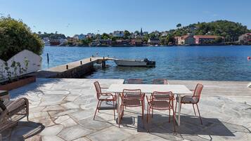 A waterfront patio with stone flooring, a table with chairs, and a view of the water with boats and buildings in the background.