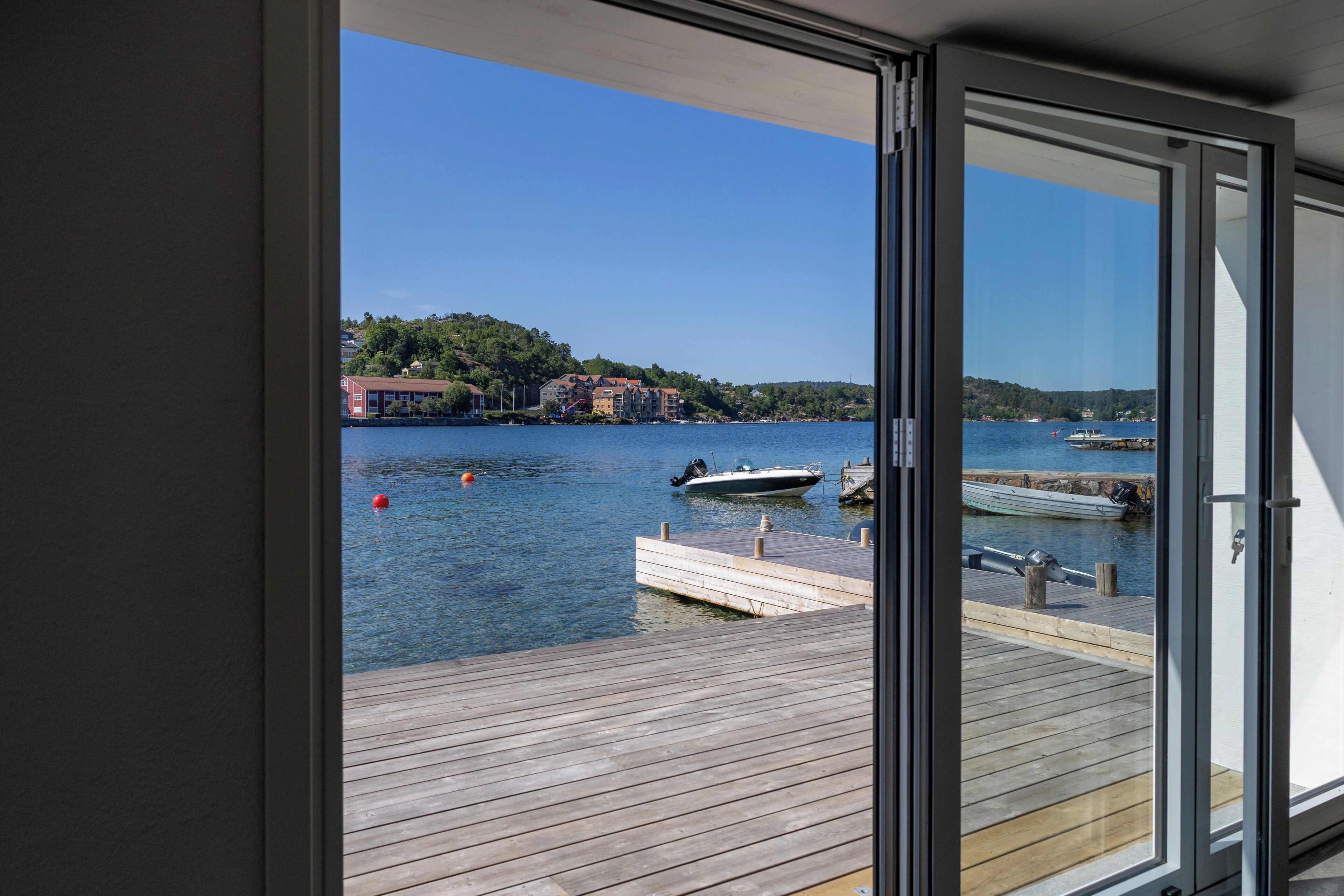 A view from a room with a large window overlooking a calm body of water, docked boats, and a distant shoreline with buildings.