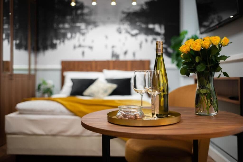 A bed with white bedding and a yellow throw, a wooden table with a bottle of wine, glasses, and a vase of yellow roses.