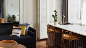 Ein modernes Wohnzimmer mit einer Couch, einem Esstisch und Stühlen, einer Küchenzeile mit Bar und einem großen Fenster mit Vorhängen.