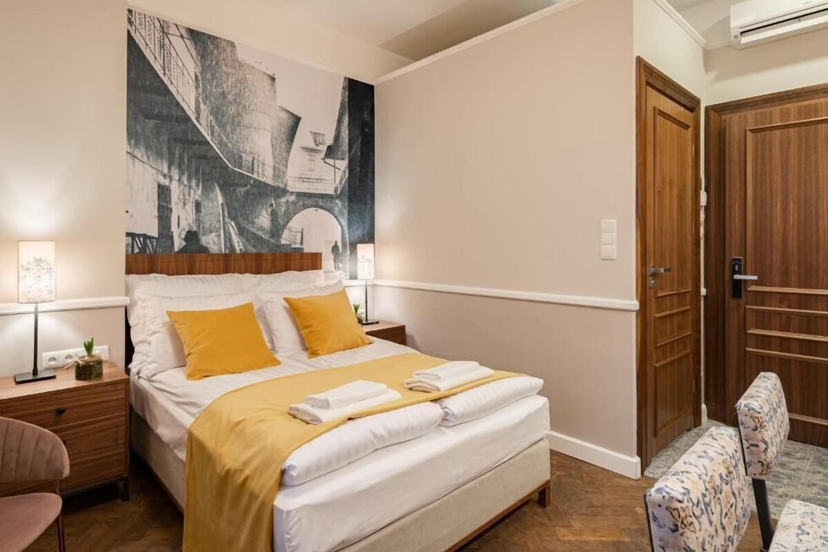 A hotel room with a bed, a nightstand, a chair, a wooden door, and a wall with a large black and white artwork.