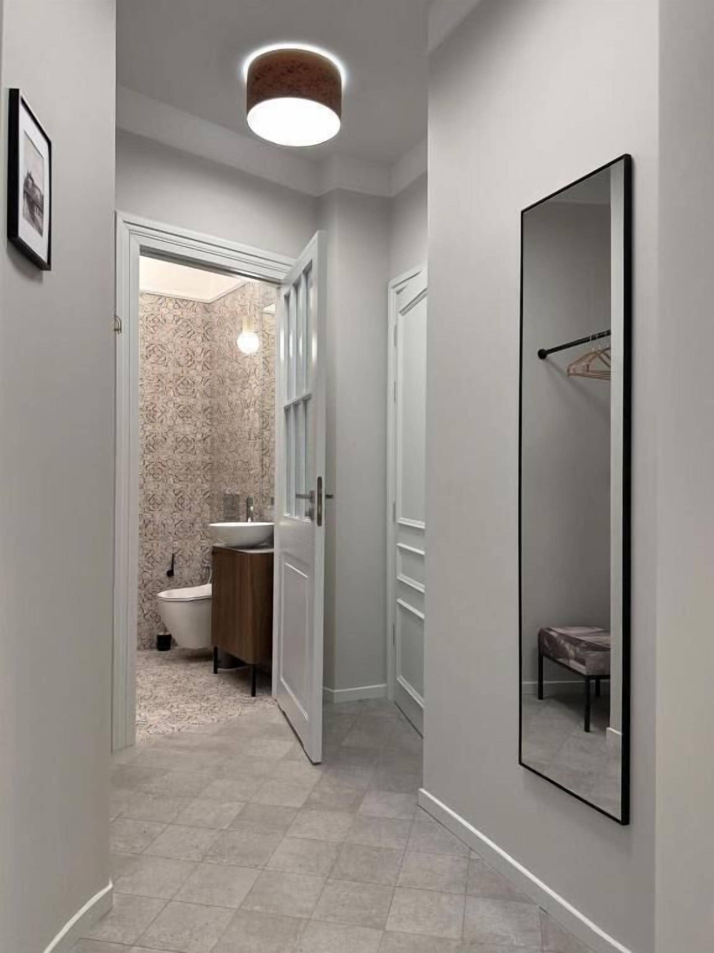Un pasillo estrecho con una puerta que da al baño, un espejo grande en la pared y una lámpara sobre ella.
