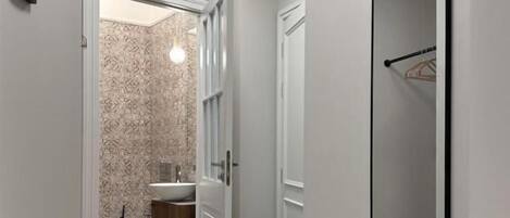 Un pasillo estrecho con una puerta que da al baño, un espejo grande en la pared y una lámpara sobre ella.