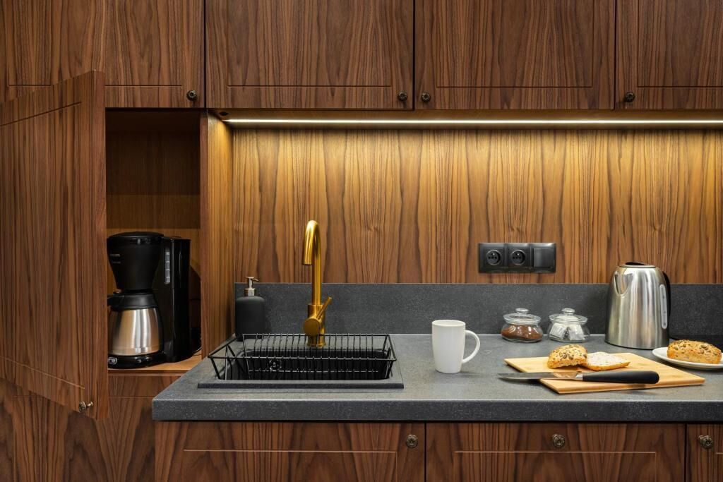 Een keuken met houten kastjes, een koffiezetapparaat, een waterkoker, een koffiekan, een mok en een snijplank met brood.