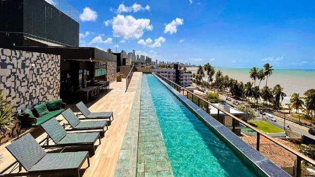 Piscina en la azotea con sillones de descanso, pared de piedra y vistas a la playa y al horizonte de la ciudad.
