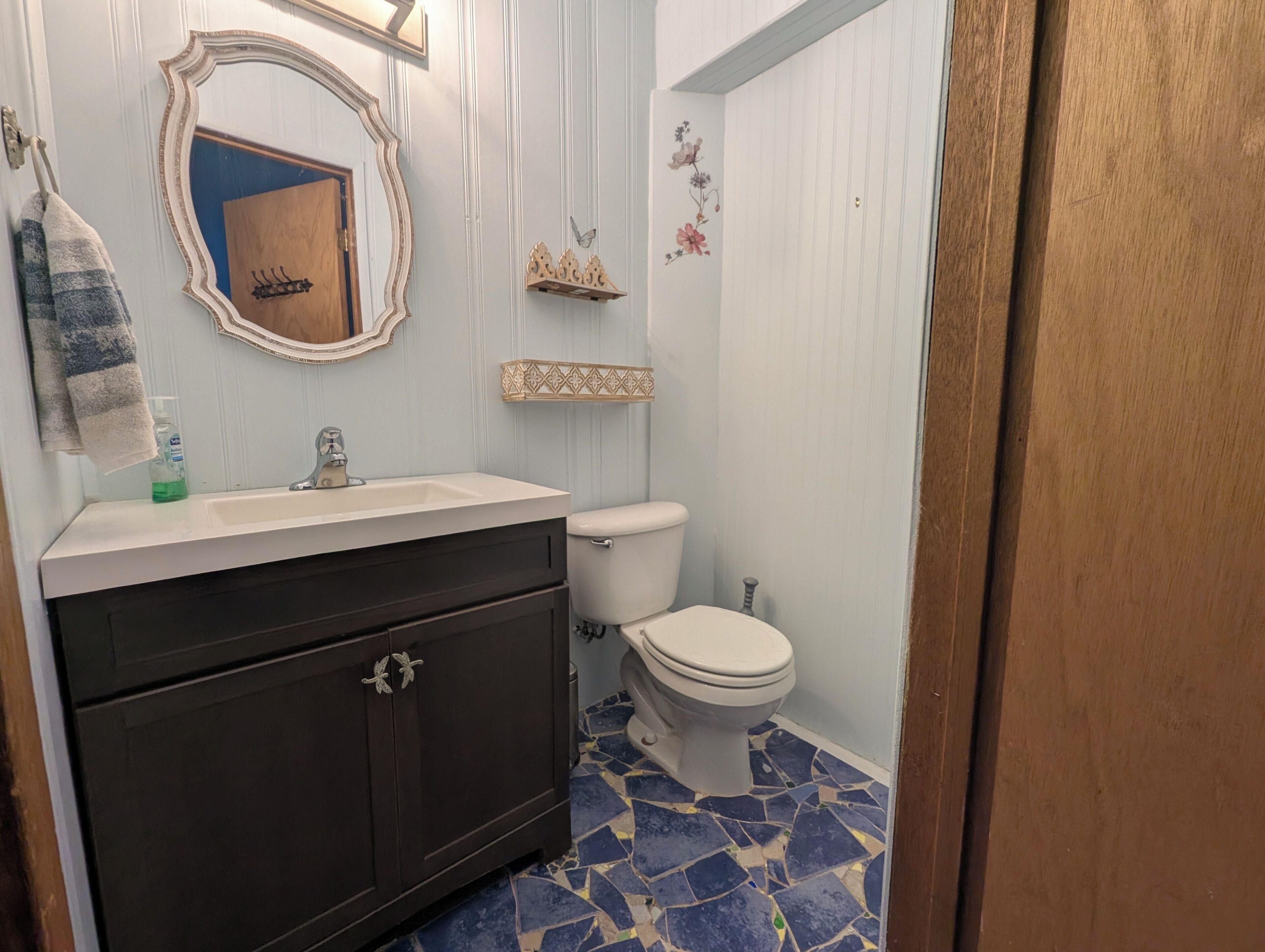 Un bagno con lavandino bianco, mobiletto in legno scuro, WC bianco, specchio decorativo e pavimento piastrellato in blu e beige.