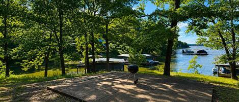 Een camping met een barbecue, omringd door bomen en een meer met boten.