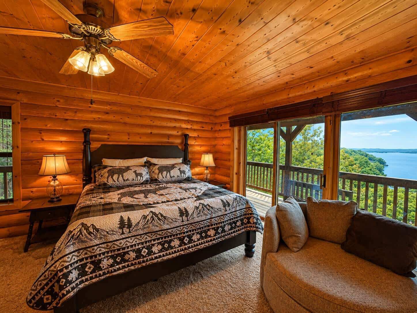 A wooden cabin bedroom with a bed, two bedside lamps, a ceiling fan, a sofa, and a large window overlooking a lake.