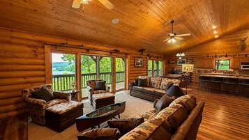 A log cabin interior with a living room, dining area, and kitchen, featuring large windows and a view of the outdoors.