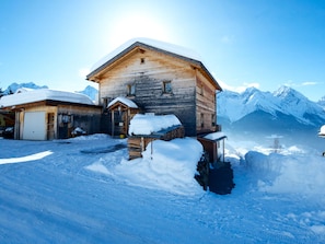 A wooden mountain cabin with a snow-covered roof, surrounded by snow-covered ground and mountains in the background.