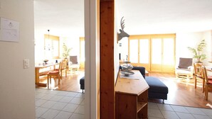 A spacious living area with a wooden bench, a dining table with chairs, and a large window with a view. - Chasa Aglia, (Ftan) by Interhome (Ftan)