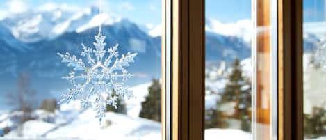 Un flocon de neige décoratif suspendu près d’une fenêtre, d’où l’on peut admirer des montagnes enneigées.