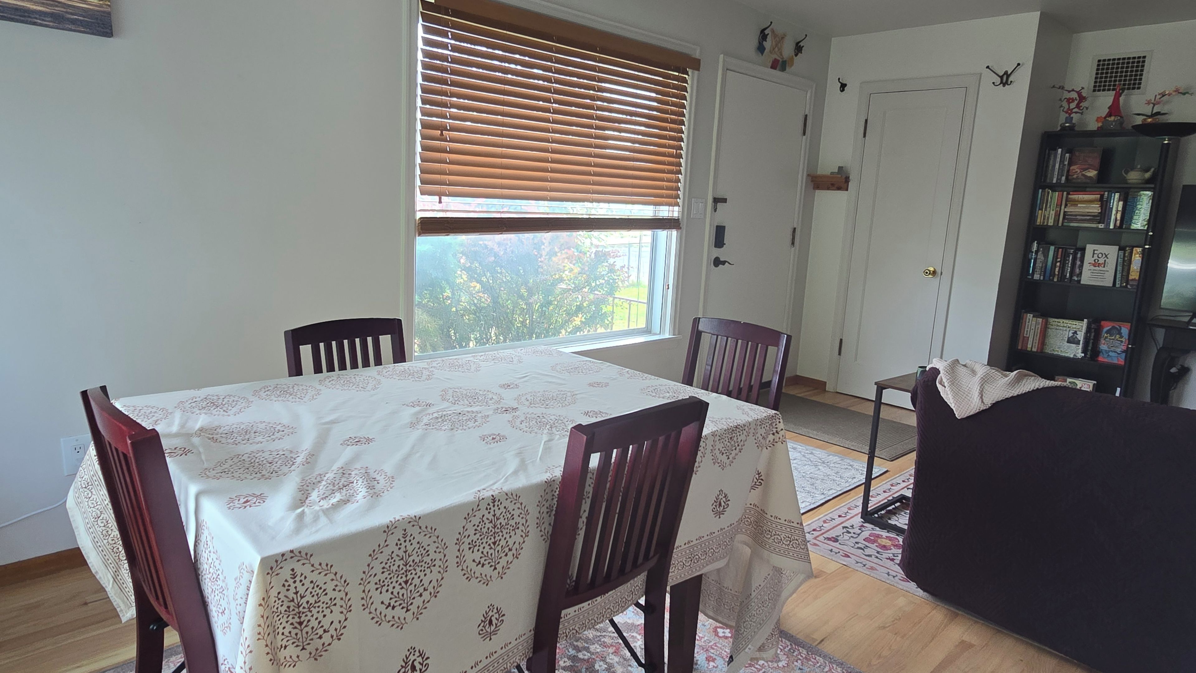 Un coin repas avec une table dressée pour quatre personnes, une fenêtre avec des stores, une bibliothèque et une porte.