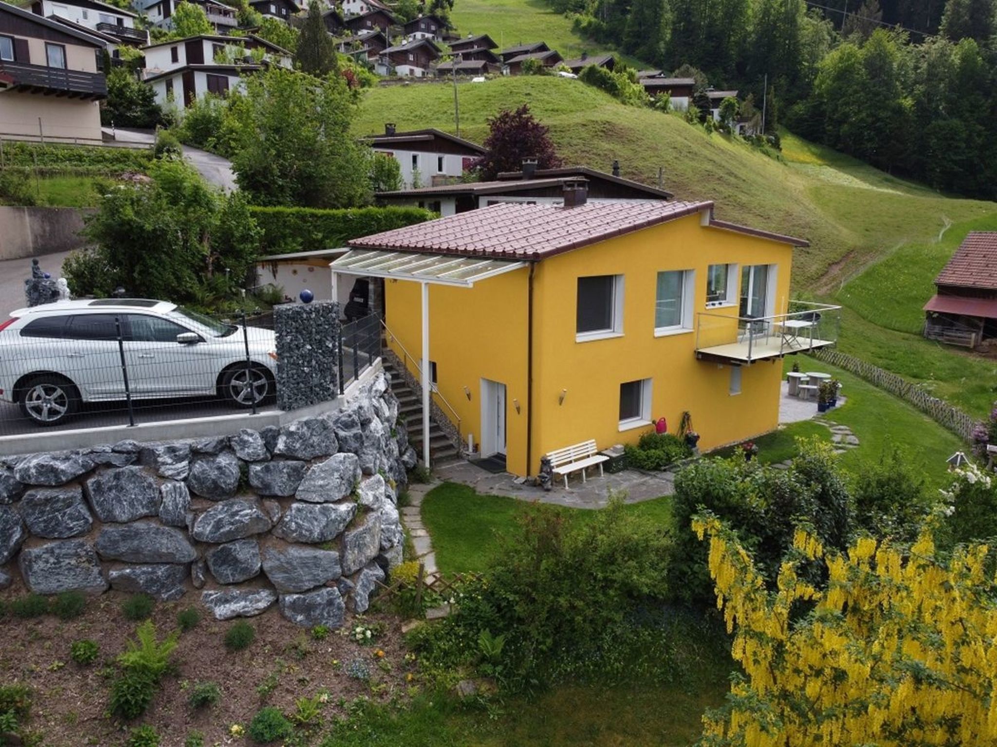 Un bâtiment jaune avec un mur en pierre, une voiture garée devant et une colline en arrière-plan.