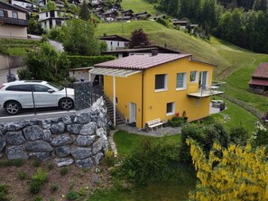 A yellow building with a stone wall, a car parked in front, and a hillside in the background.