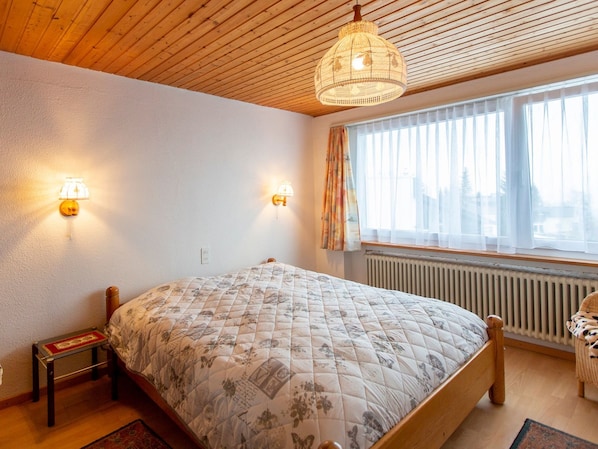 A bedroom with a wooden ceiling, a bed with a floral quilt, two wall-mounted lamps, a window with curtains, and a radiator.
