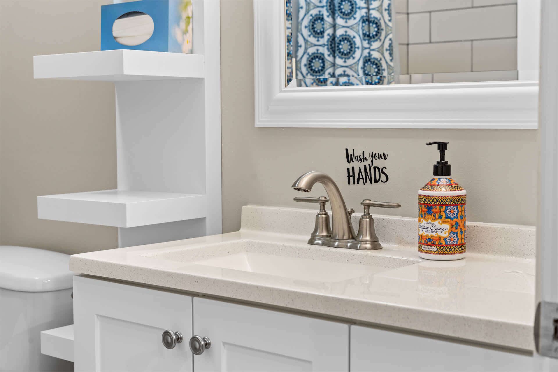A bathroom with a white sink, a mirror, a decorative hand soap dispenser, and a 'Wash your hands' sign.