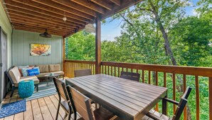 Eine überdachte Terrasse mit Holztisch und -stühlen, einer Couch mit Kissen und Blick auf Bäume.