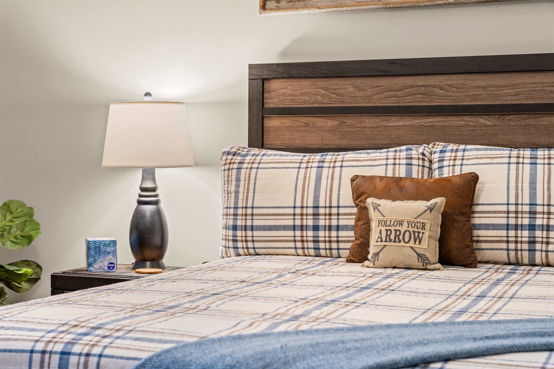 Una cama bien hecha con ropa de cama escocesa, un cabecero de madera, una lámpara de noche y una almohada con una frase motivacional.