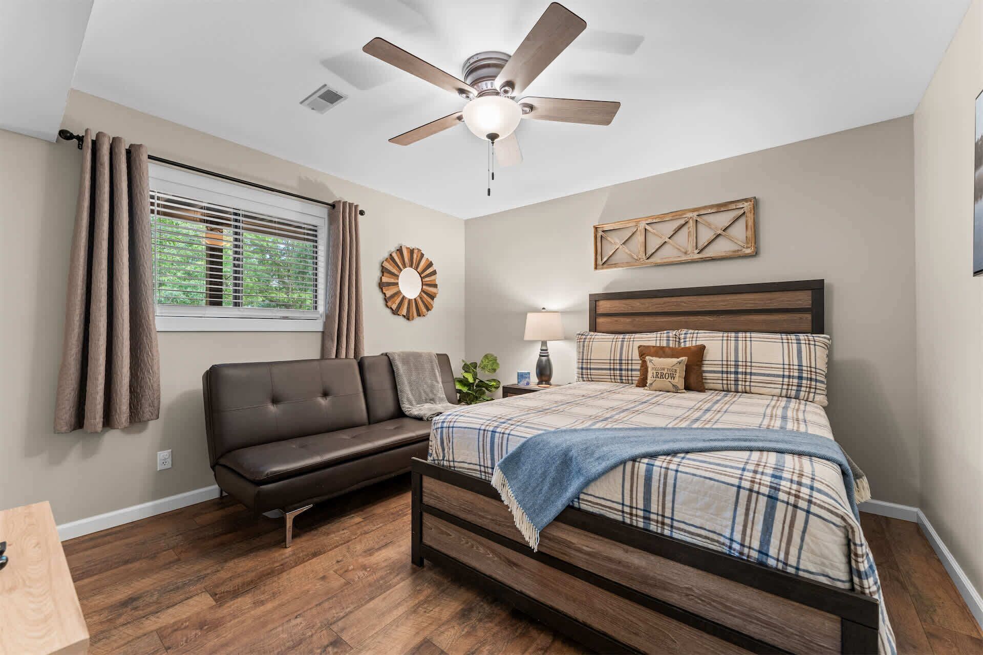 A bedroom with a bed, a ceiling fan, a window with curtains, a wall clock, and a bedside table with a lamp.