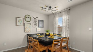 Une salle à manger avec une table noire, des chaises en bois, un lustre, des œuvres d’art encadrées et une fenêtre ornée de rideaux à motifs.