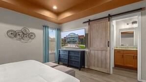 Une chambre à coucher comprenant un lit, une commode, une porte coulissante, un téléviseur affichant une image d’un bâtiment, et une salle de bain visible à travers la porte.